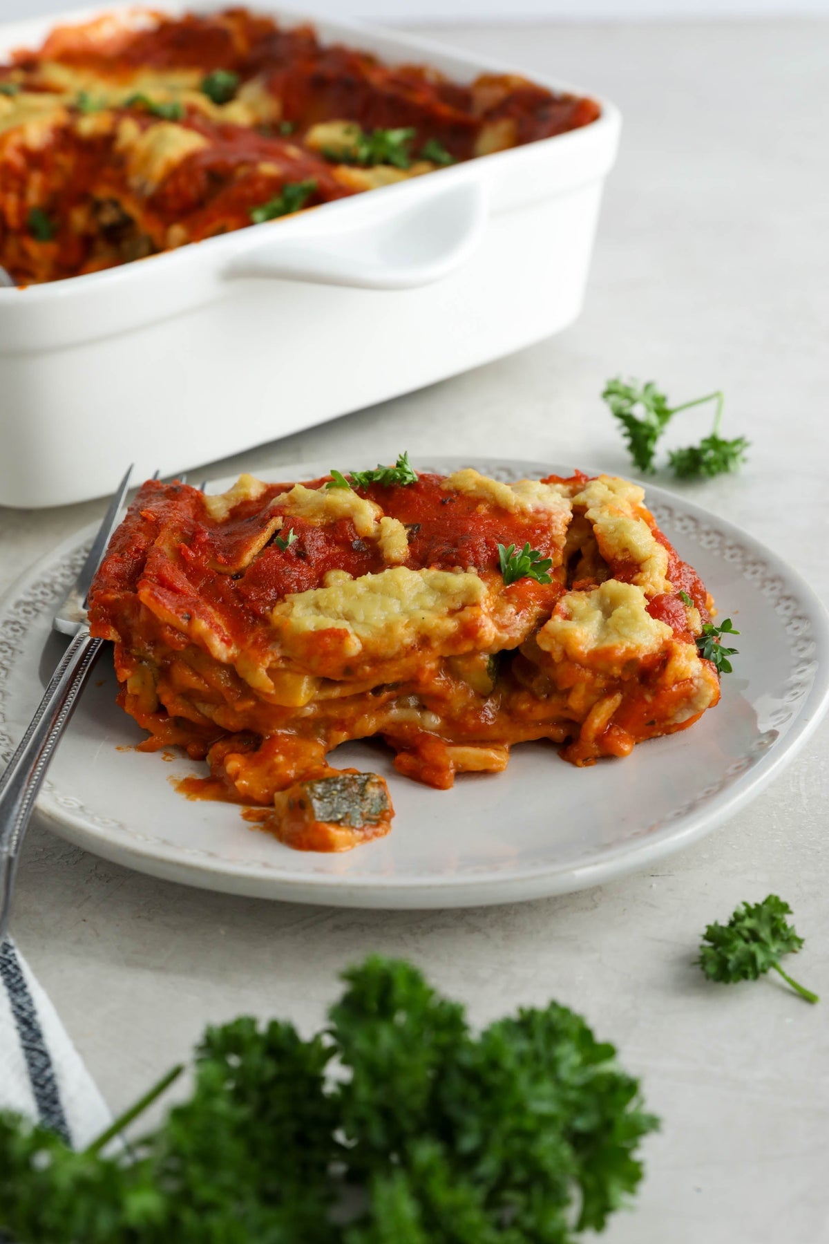Slice of lasagna on a white plate with a casserole dish of lasagna in the background