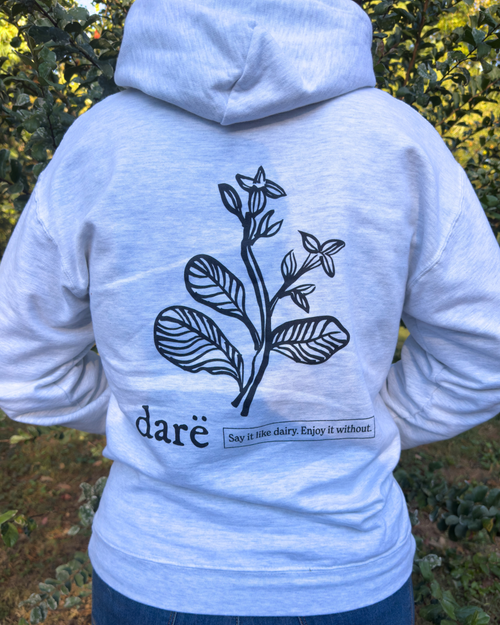 Load image 1 in gallery view. A person viewed from behind wearing a light heather gray hoodie with a black botanical line drawing and the words Say it like dairy. Enjoy it without. printed on the back, standing outdoors in front of green foliage.