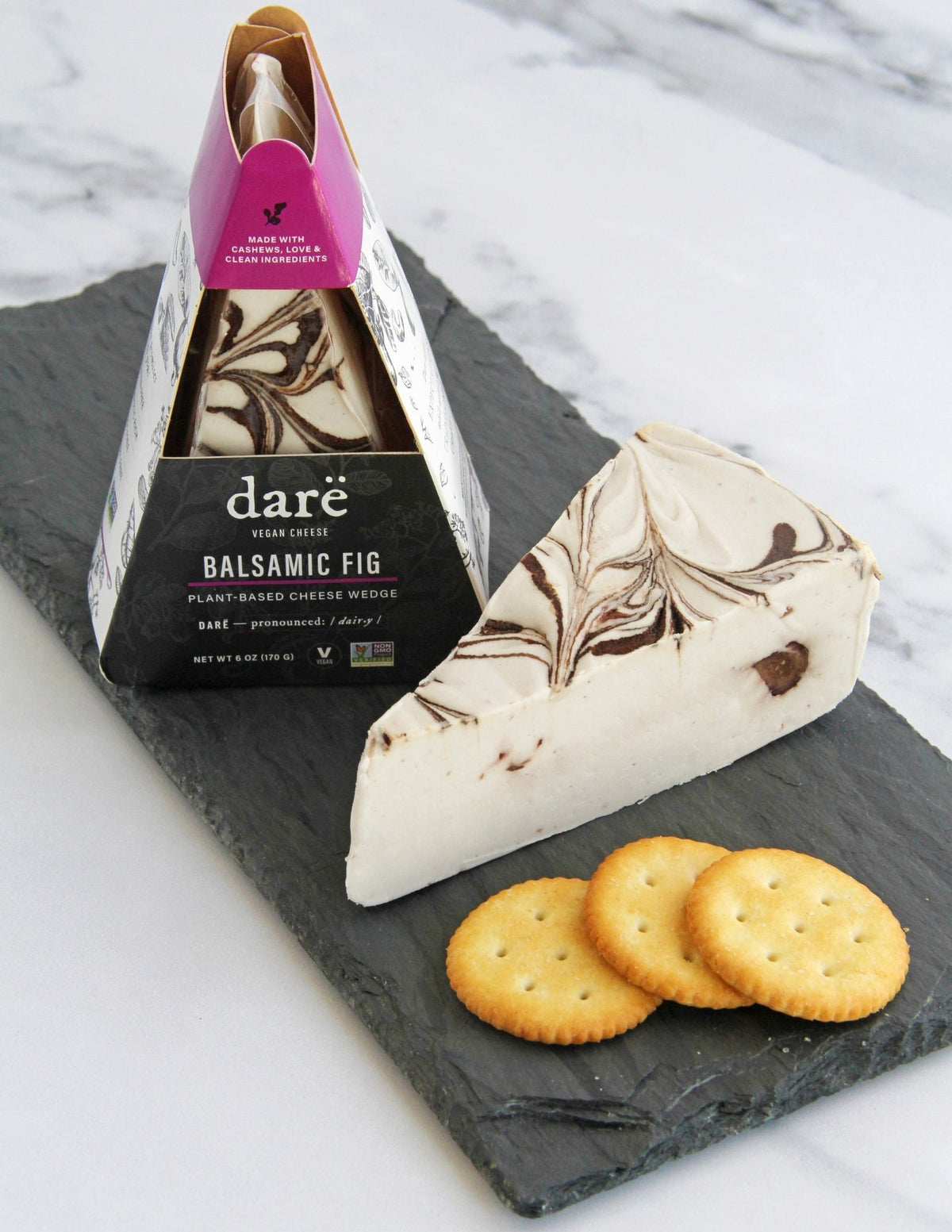 A wedge of Darë Vegan Cheese Balsamic Fig plant-based cheese on a slate board with crackers, with the packaged product standing behind it on a light marble surface.