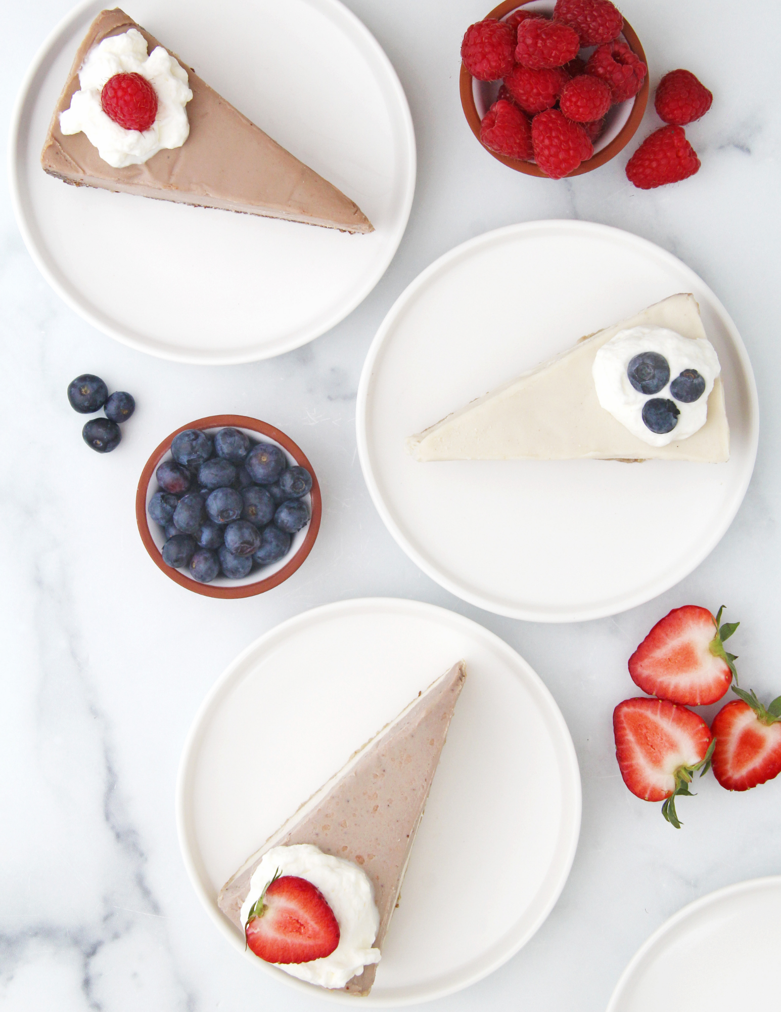 Three slices of vegan cheesecake on white plates, styled with fresh berries and strawberries on a white marble surface. The top-left slice is chocolate cake with whipped cream and a raspberry, the middle-right slice original cheesecake with blueberries, and the bottom-left slice is strawberry cheesecake with whipped cream and a strawberry.