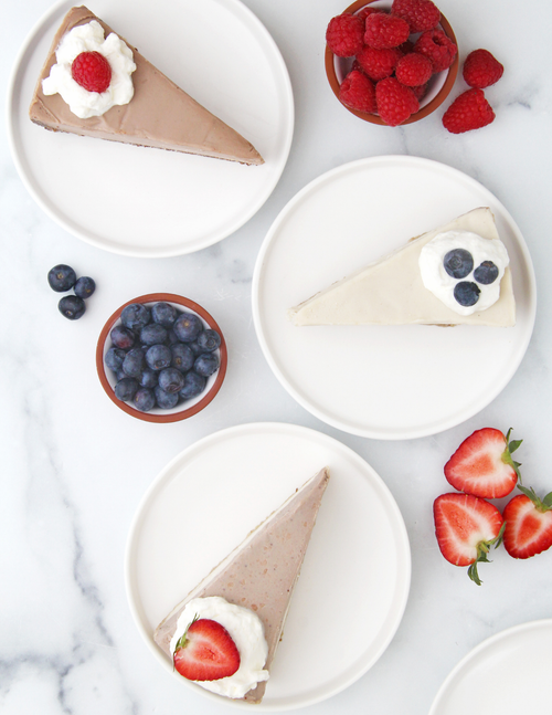 Load image 1 in gallery view. Three slices of vegan cheesecake on white plates, styled with fresh berries and strawberries on a white marble surface. The top-left slice is chocolate cake with whipped cream and a raspberry, the middle-right slice original cheesecake with blueberries, and the bottom-left slice is strawberry cheesecake with whipped cream and a strawberry.