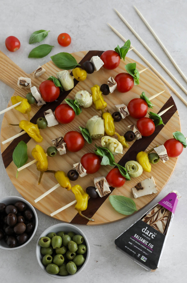 Wooden board with skewers of vegetables and cheese, surrounded by olives and a package of Dare Balsamic Fig on a light gray background.