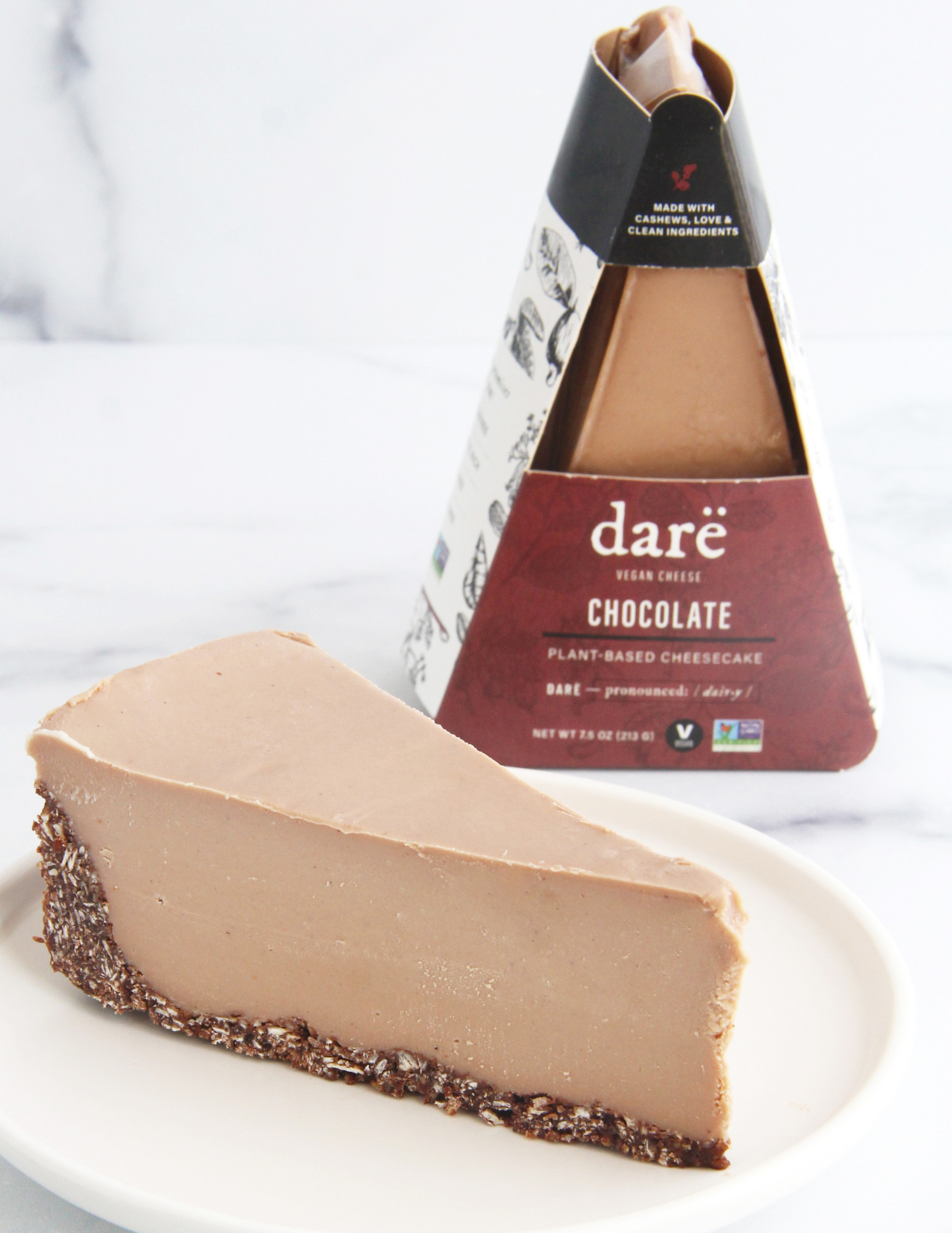 A slice of daré plant-based chocolate cheesecake on a white plate, with the branded triangular package standing behind it on a light marble surface.