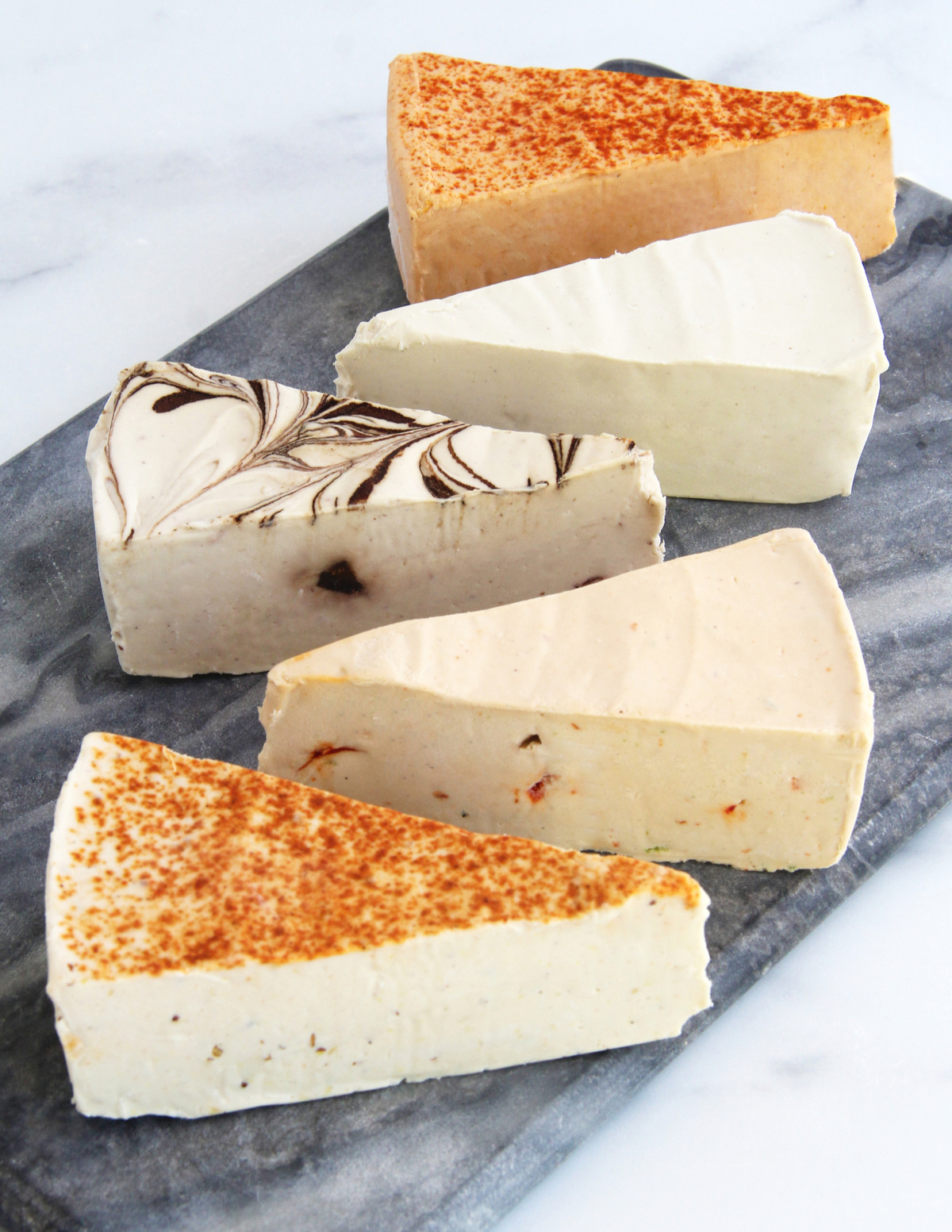 Four triangular wedges of artisanal vegan cheese displayed on a dark slate serving board over a white marble surface