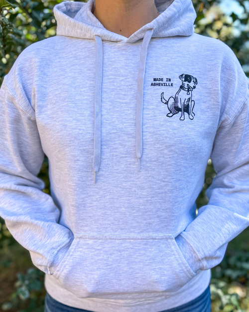 Load image 1 in gallery view. A person wearing a light heather gray pullover hoodie with a black graphic of a dog and the text 'MADE IN ASHEVILLE' on the left chest, photographed outdoors with blurred green foliage in the background.