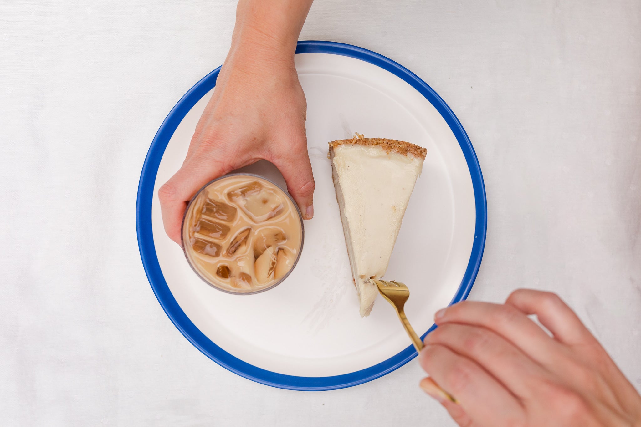 Person holding a cup of iced coffee and a slice of cake with a gold fork on a white background.