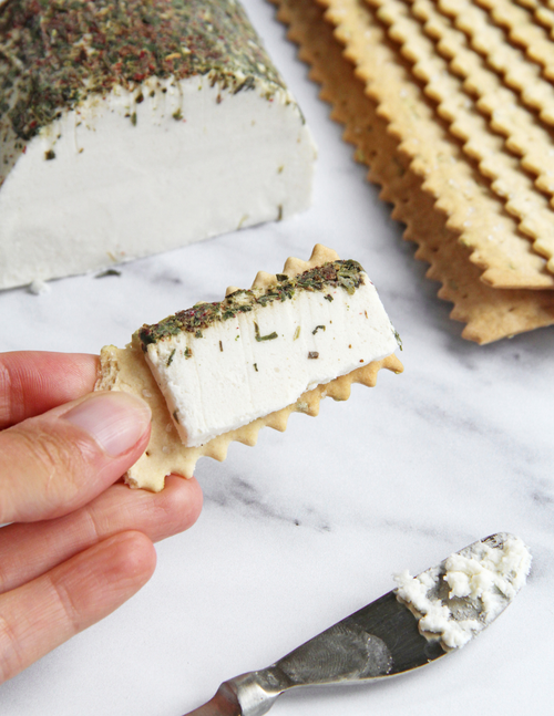Load image 1 in gallery view. A hand holds a square cracker topped with a thick slice of vegan goat cheese spread with herbs, with a large block of the cheese and a stack of crackers in the background on a white marble surface.
