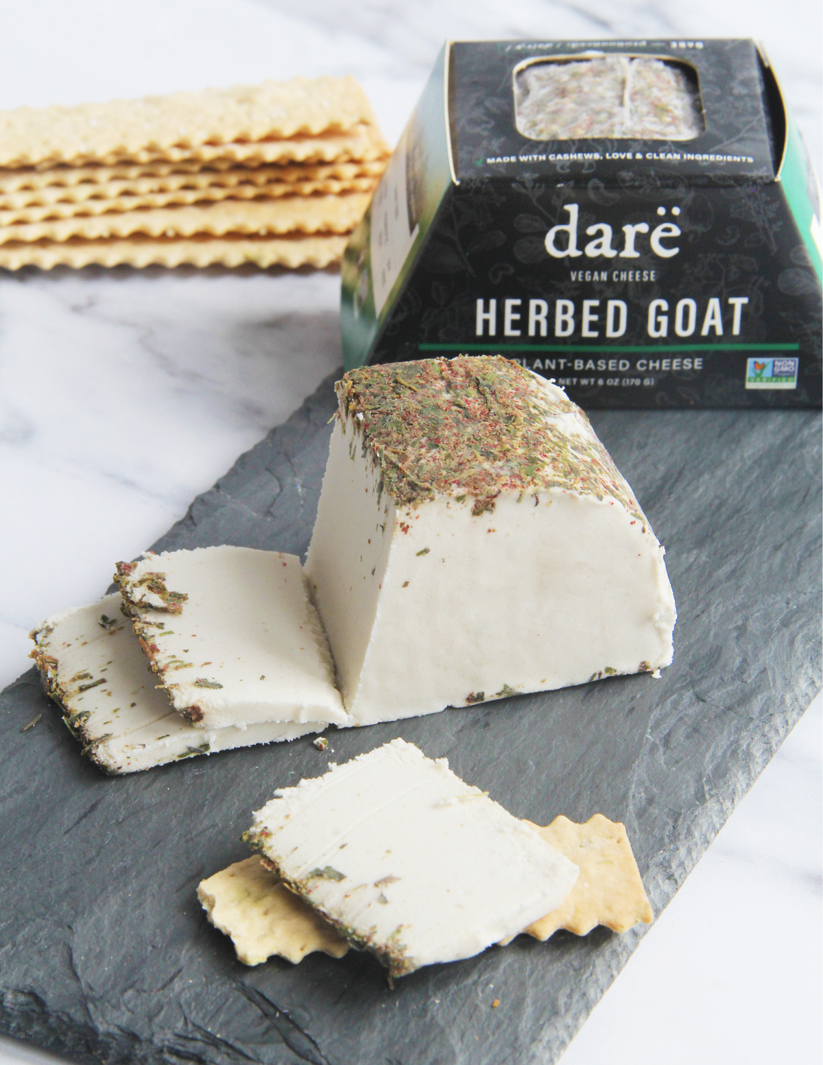 A wedge and slices of Dare Herbed Goat Vegan Cheese displayed on a dark slate board with crackers, alongside its black packaging box on a white marble surface.