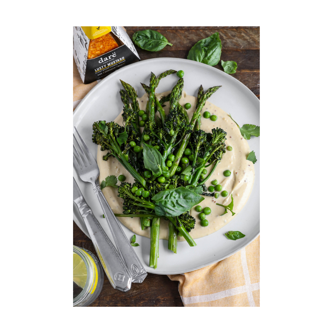 Green vegetables over a bed of vegan lusty mustard cheese sauce on a white plate with a silver fork and knife on the side.