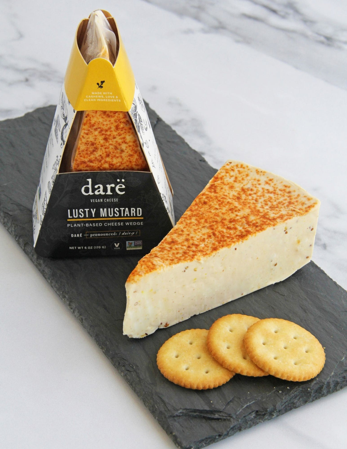 A slice of Darë Vegan Cheese Lusty Mustard plant-based cheese on a slate board with crackers, with the packaged product standing behind it on a light marble surface.