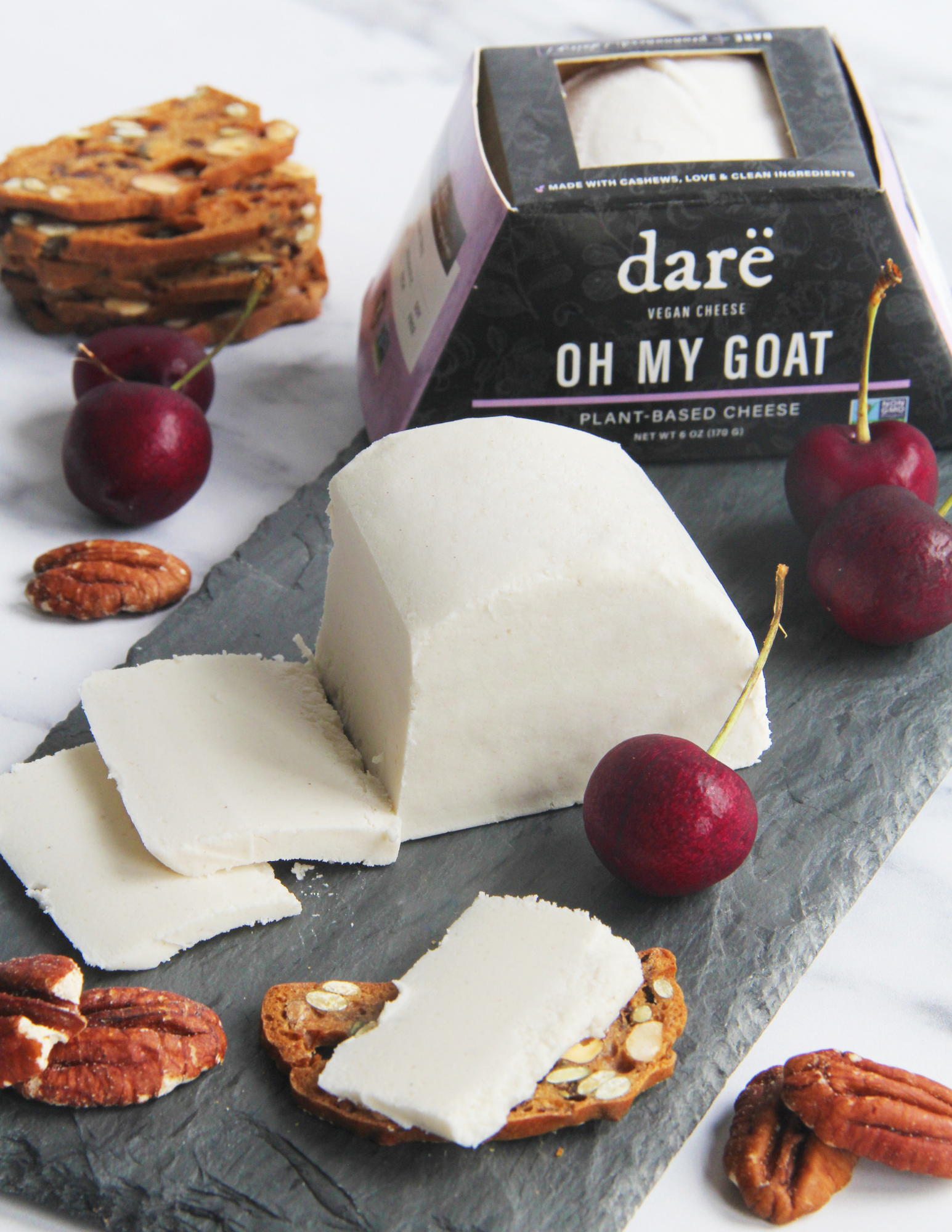 A wedge and slices of Dare Vegan Cheese Plant-Based Cheese 'Oh My Goat' displayed on a dark slate board with cherries, pecans, and almond crackers, with the product packaging in the background.