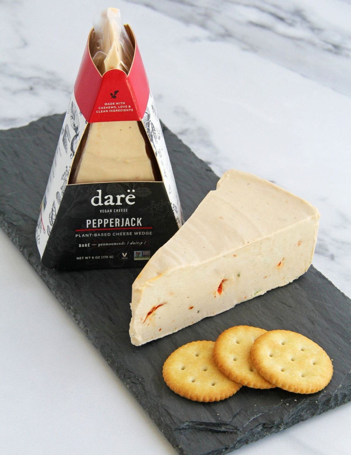 A slice of Darë Vegan Cheese Pepperjack plant-based cheese on a slate board with crackers, with the packaged product standing behind it on a light marble surface.