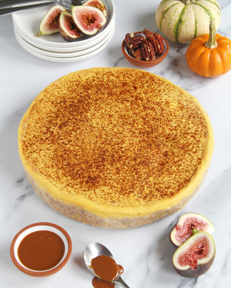 A vegan pumpkin cheesecake, presented on a white marble surface with seasonal garnishes including figs, pecans, and small pumpkins.