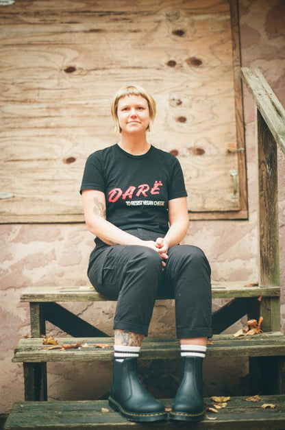 woman sitting on wooden steps with black tshirt on. red lettering reads dare white lettering reads to resist vegan cheese