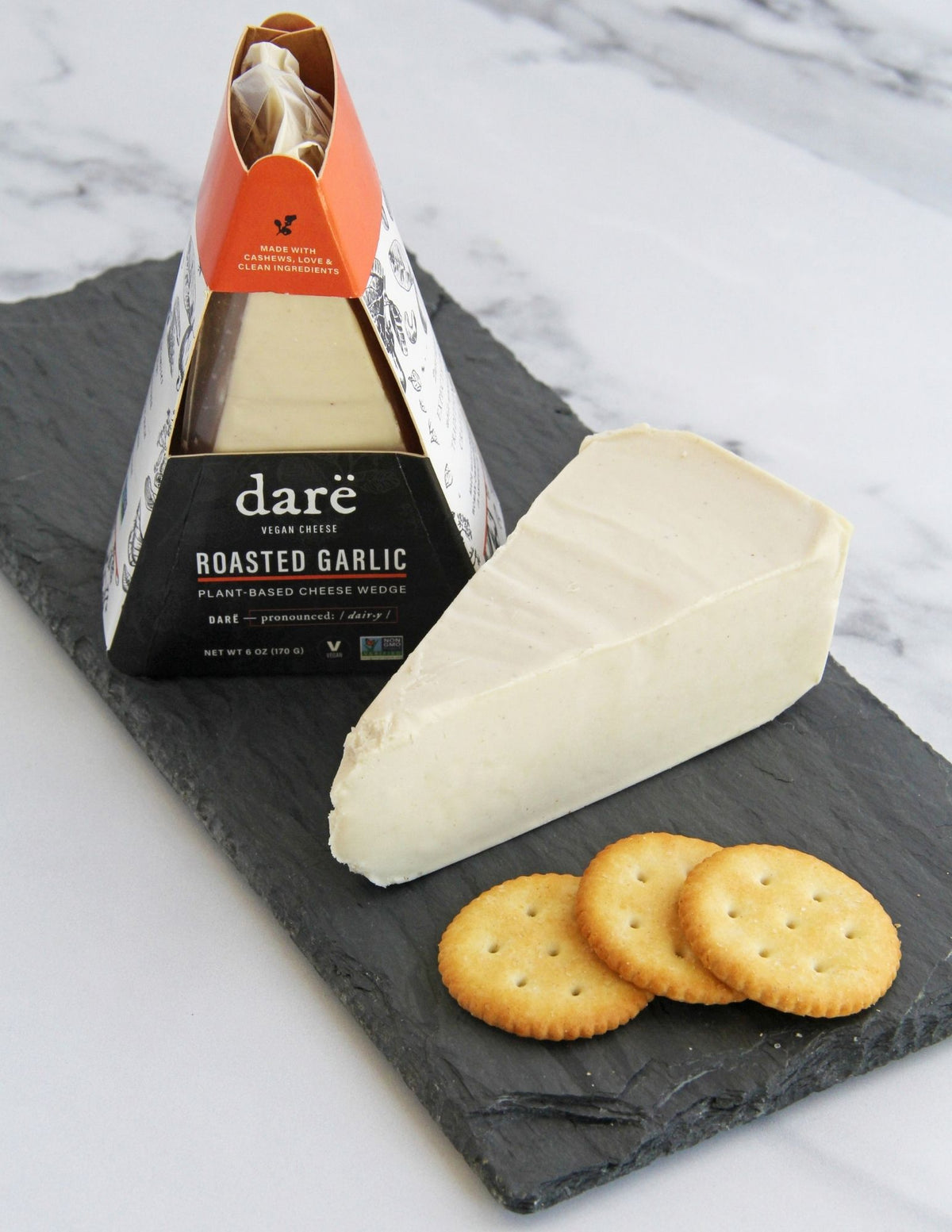 A slice of dare vegan cheese Roasted Garlic plant-based cheese on a slate board with crackers, with the packaged product standing behind it on a light marble surface.