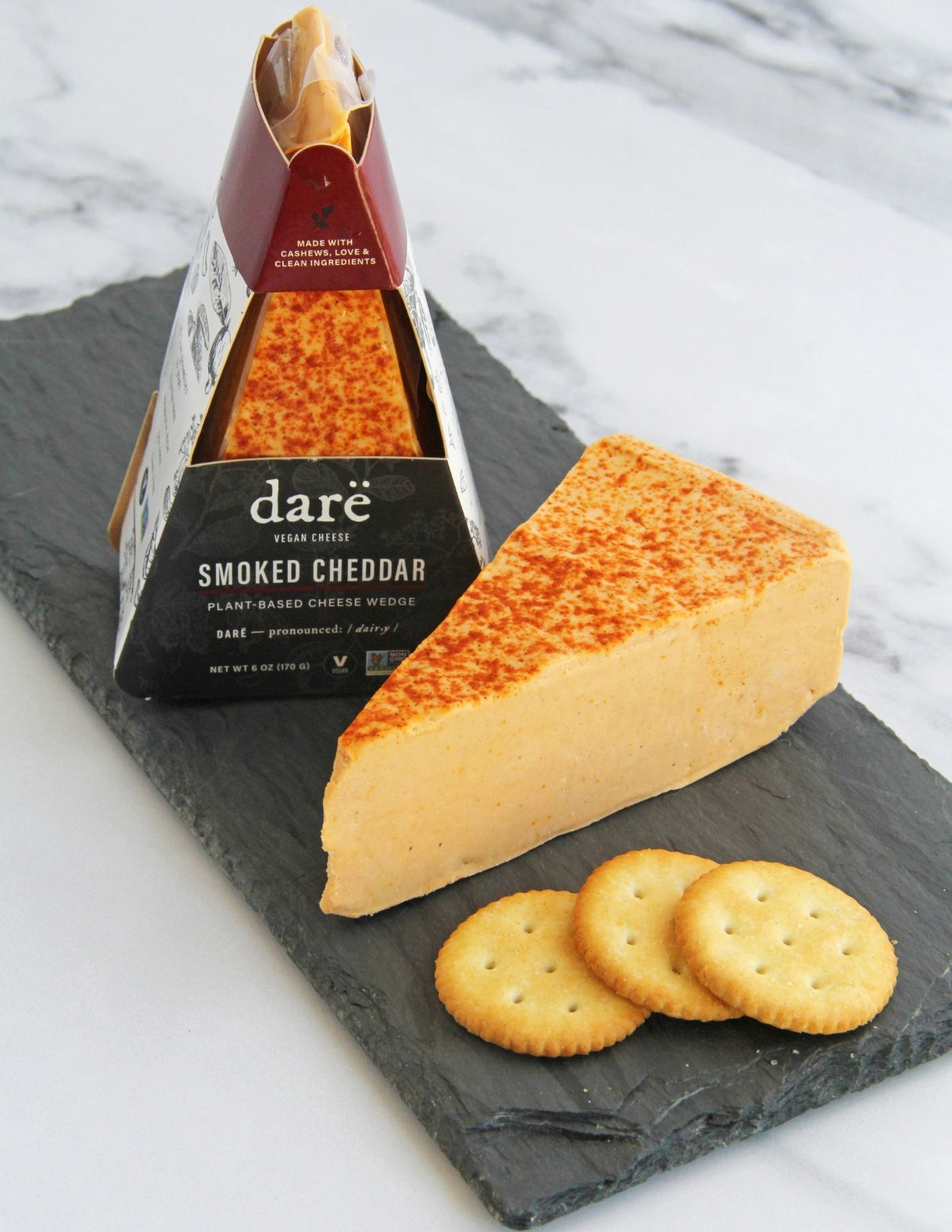 A wedge of Darë Vegan Smoked Cheddar plant-based cheese on a slate board with crackers, with the packaged product standing behind it on a light marble surface.