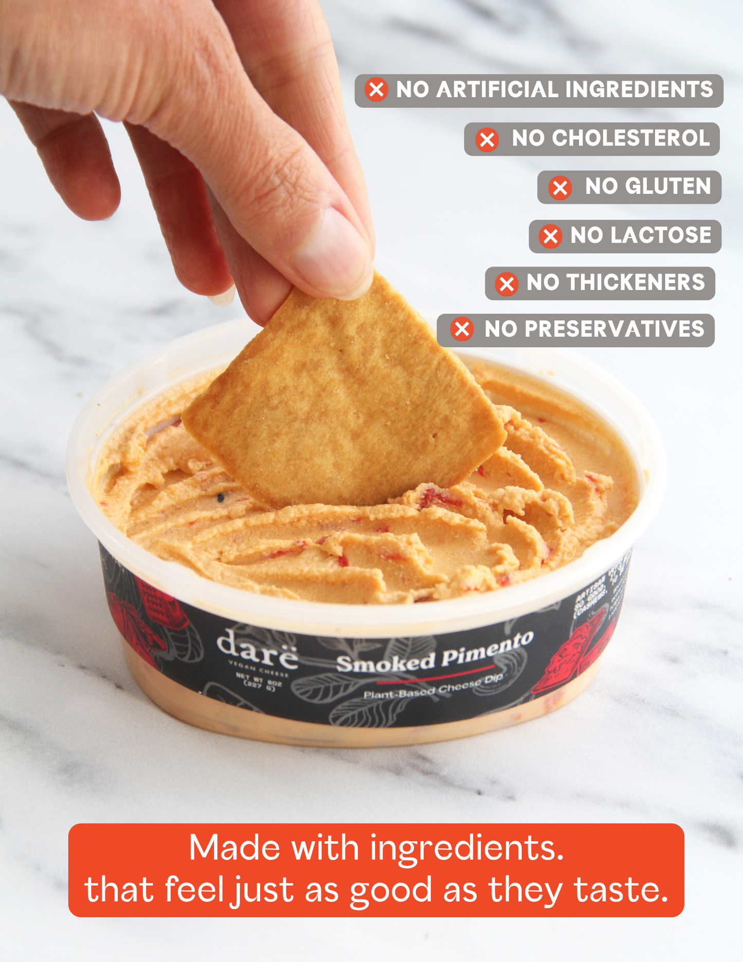 A hand dipping a pita chip into a container of dare Smoked Pimento Plant-Based Cheese Dip, set on a white marble background with text overlays highlighting product benefits.