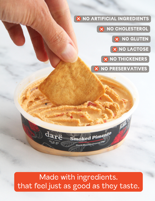 Load image 1 in gallery view. A hand dipping a pita chip into a container of dare Smoked Pimento Plant-Based Cheese Dip, set on a white marble background with text overlays highlighting product benefits.