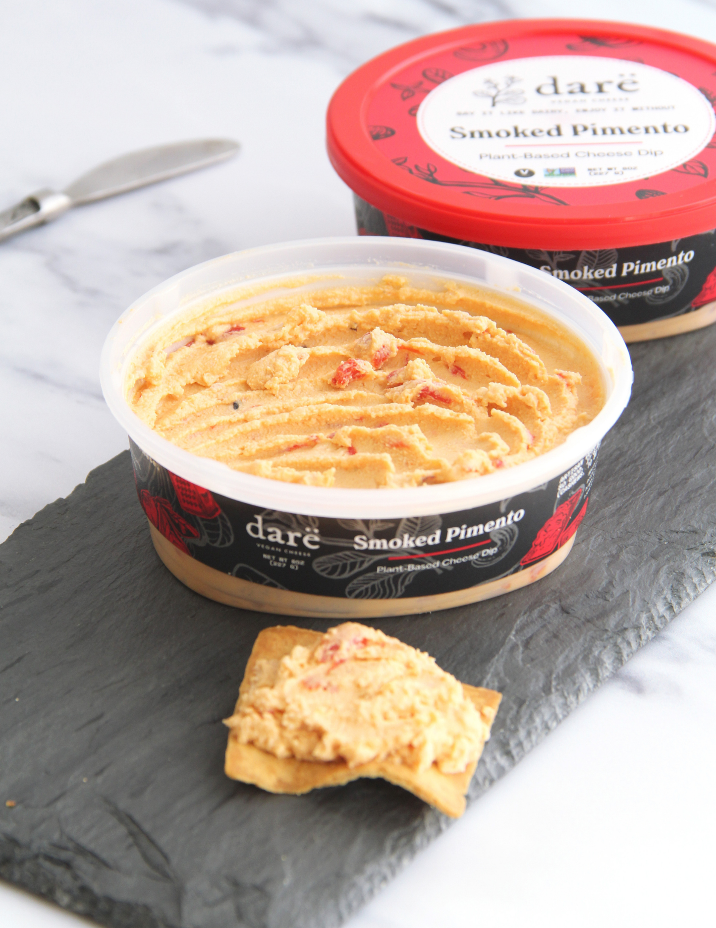 A tub of daré Smoked Pimento cheese spread in the foreground with a small cracker topped with spread, and a second tub with a red lid in the background on a dark slate board over a white marble surface.