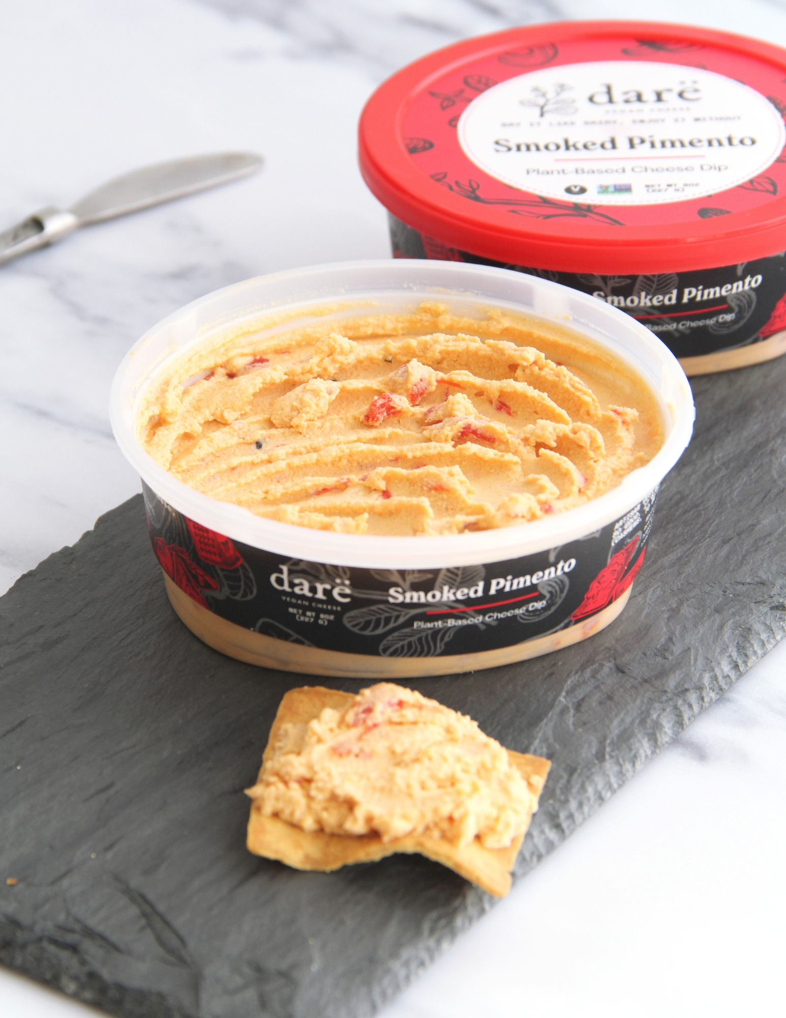 A tub of daré Smoked Pimento cheese spread in the foreground with a small cracker topped with spread, and a second tub with a red lid in the background on a dark slate board over a white marble surface.