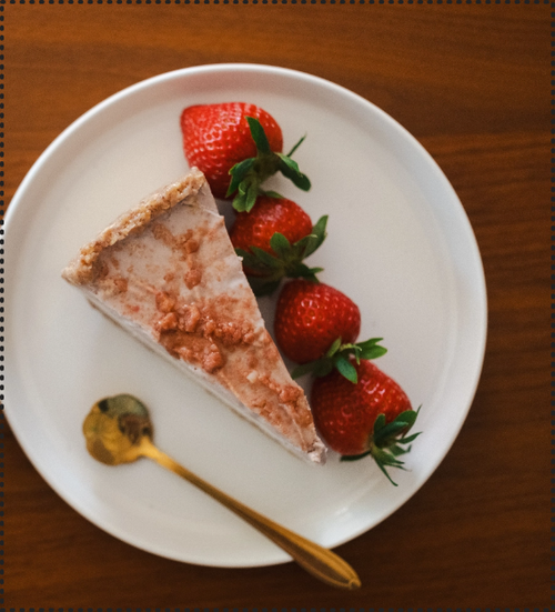 Load image 1 in gallery view. Sliced of vegan strawberry shortcake cheesecake on a white plate lined with four whole strawberries with a gold spoon with a wooden background