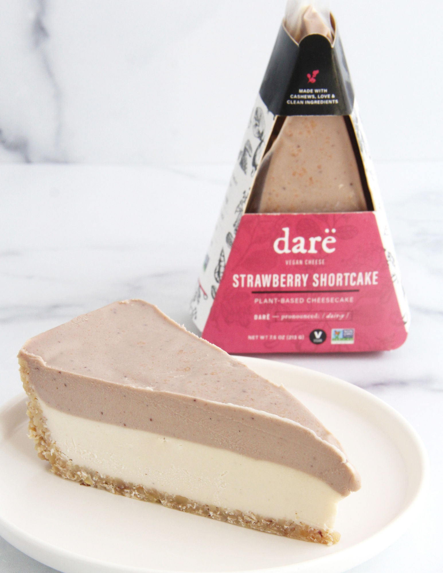 A slice of dare vegan cheese Strawberry Shortcake plant-based cheesecake on a white plate, with the packaged product standing behind it on a light marble surface.
