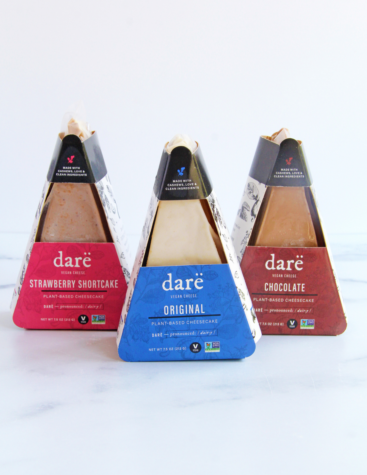 Three packaged daré plant-based cheesecakes arranged in a row on a light marble surface against a white background. The left cake is strawberry shortcake with a pink label, the middle cake is original with a blue label, and the right cake is chocolate with a brown label.