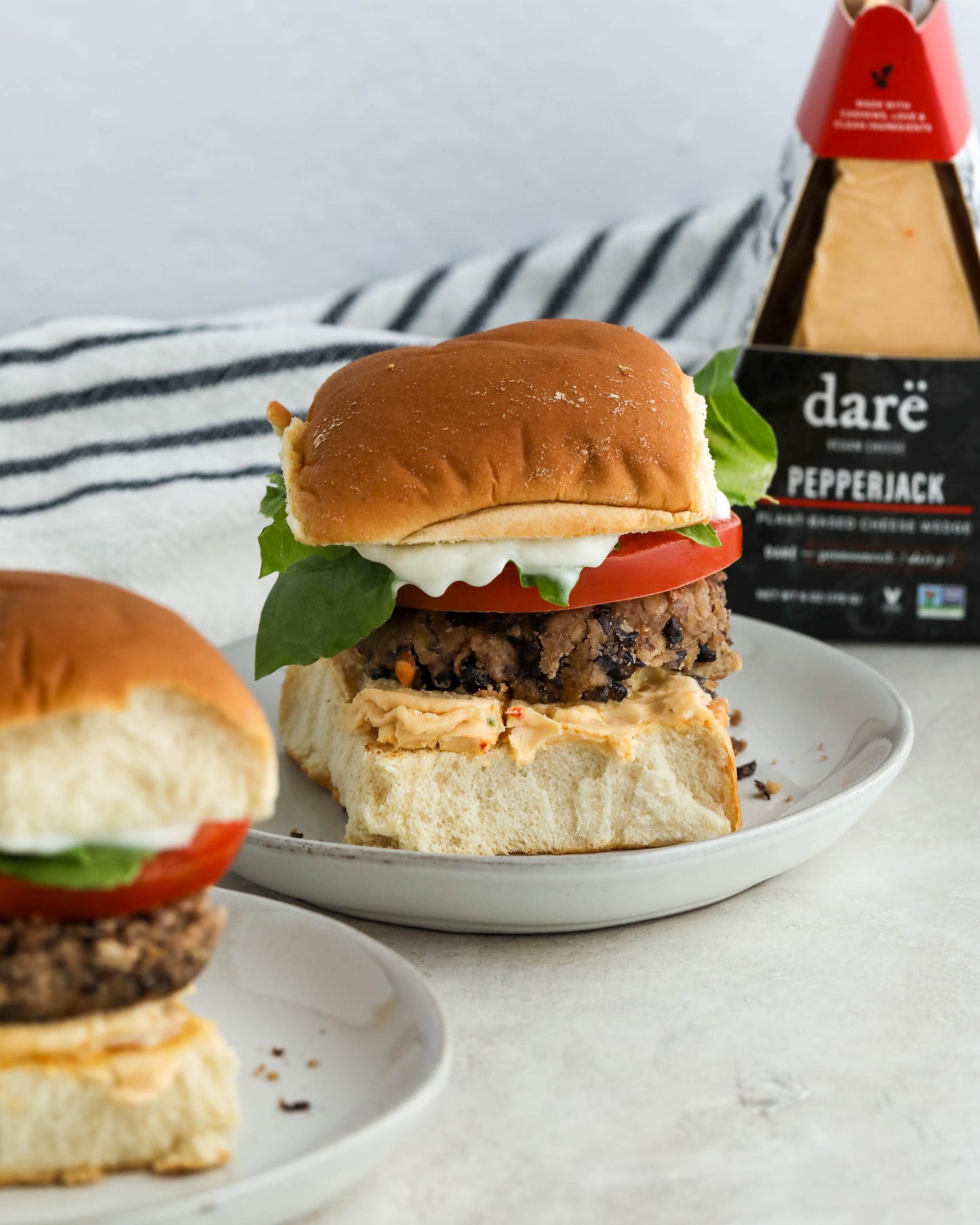 Two burgers on buns with lettuce, tomato, and cheese, with a wedge of Dare Pepperjack cheese in the background.