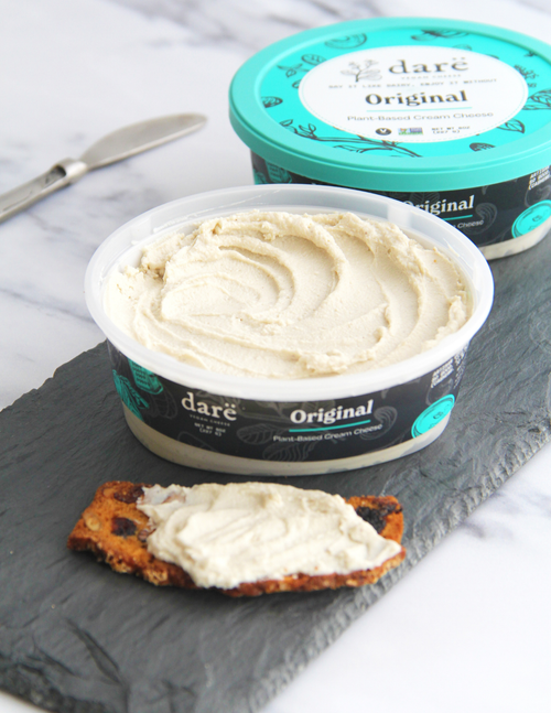 Load image 1 in gallery view. An open tub of dare vegan cheese Original Cream Cheese sits on a dark slate board with a slice of toast spread with the cream cheese, alongside a closed container and a knife on a white marble surface.