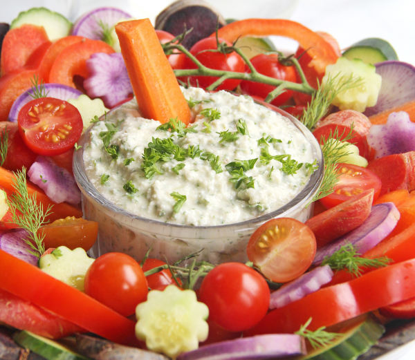 A clear glass bowl filled with a vegan ranch dip, garnished with chopped green herbs, and surrounded by sliced tomatoes, red bell peppers, cucumbers, red onions, and a carrot on a bed of mixed vegetables.
