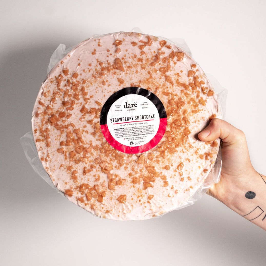 whole strawberry shortcake cheesecake round, being held by a human hand. white background, and cheesecake has pink strawberry bits on top, all wrapped in plastic packaging