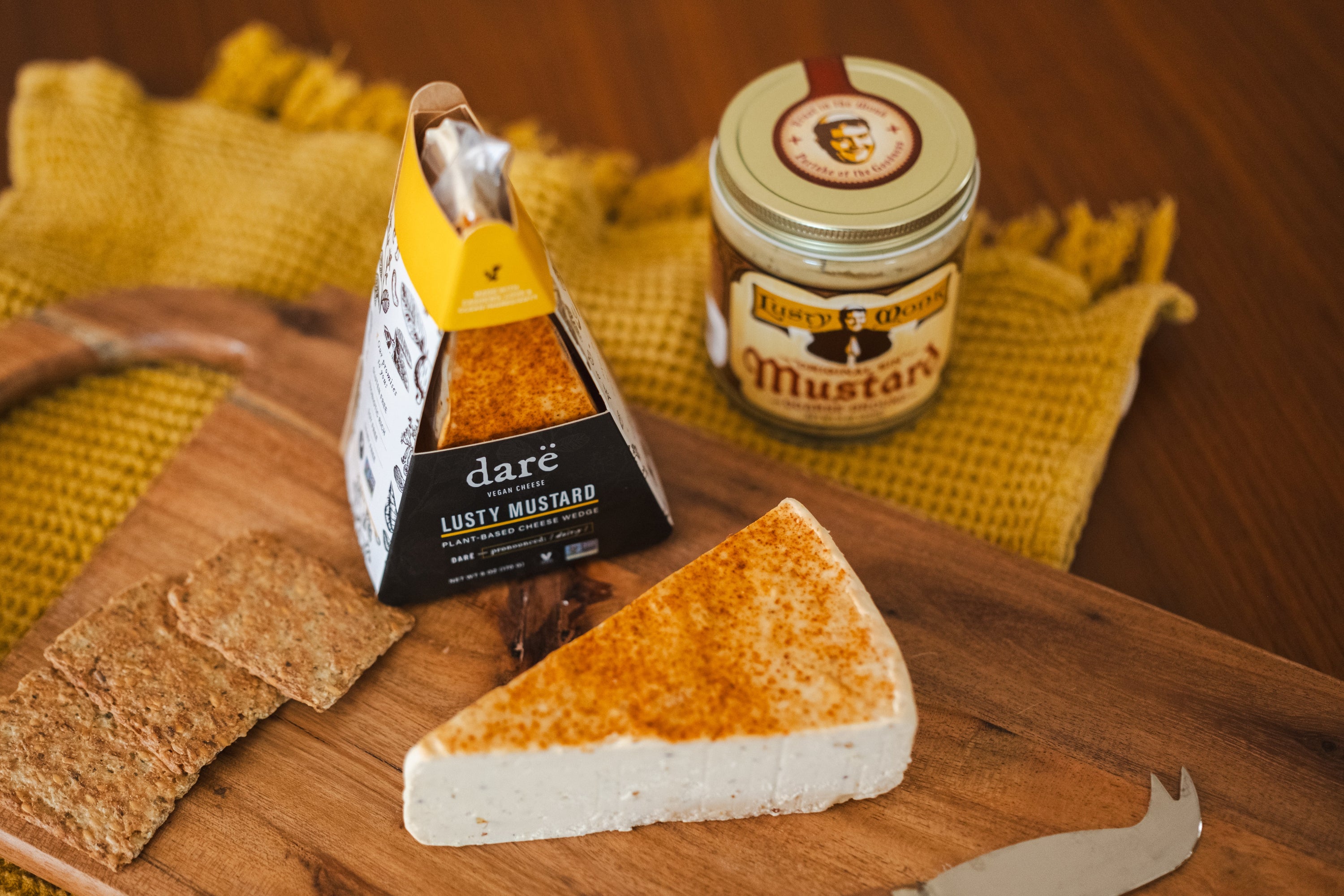 Darë Lusty Mustard Wedge packaging next to Lusty Monk Mustard container. Lusty Mustard Wedge on wooden cheeseboard with crackers.