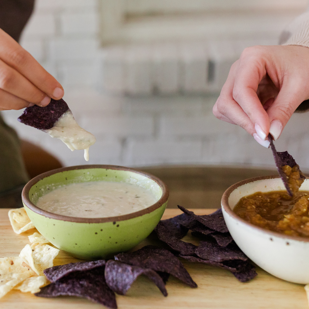 Queso Blanco cheese dip in ceramic dishes. hands using blue corn tortilla chips to dip into vegan cheese.