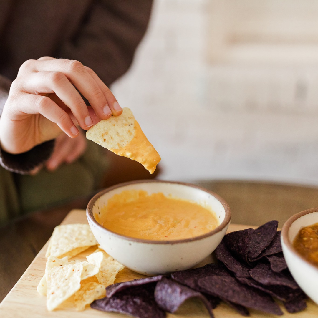 Hand holding tortilla chip dips into Darë Vegan Cheese Spicy Queso. white and blue corn tortilla chips. queso in ceramic dish