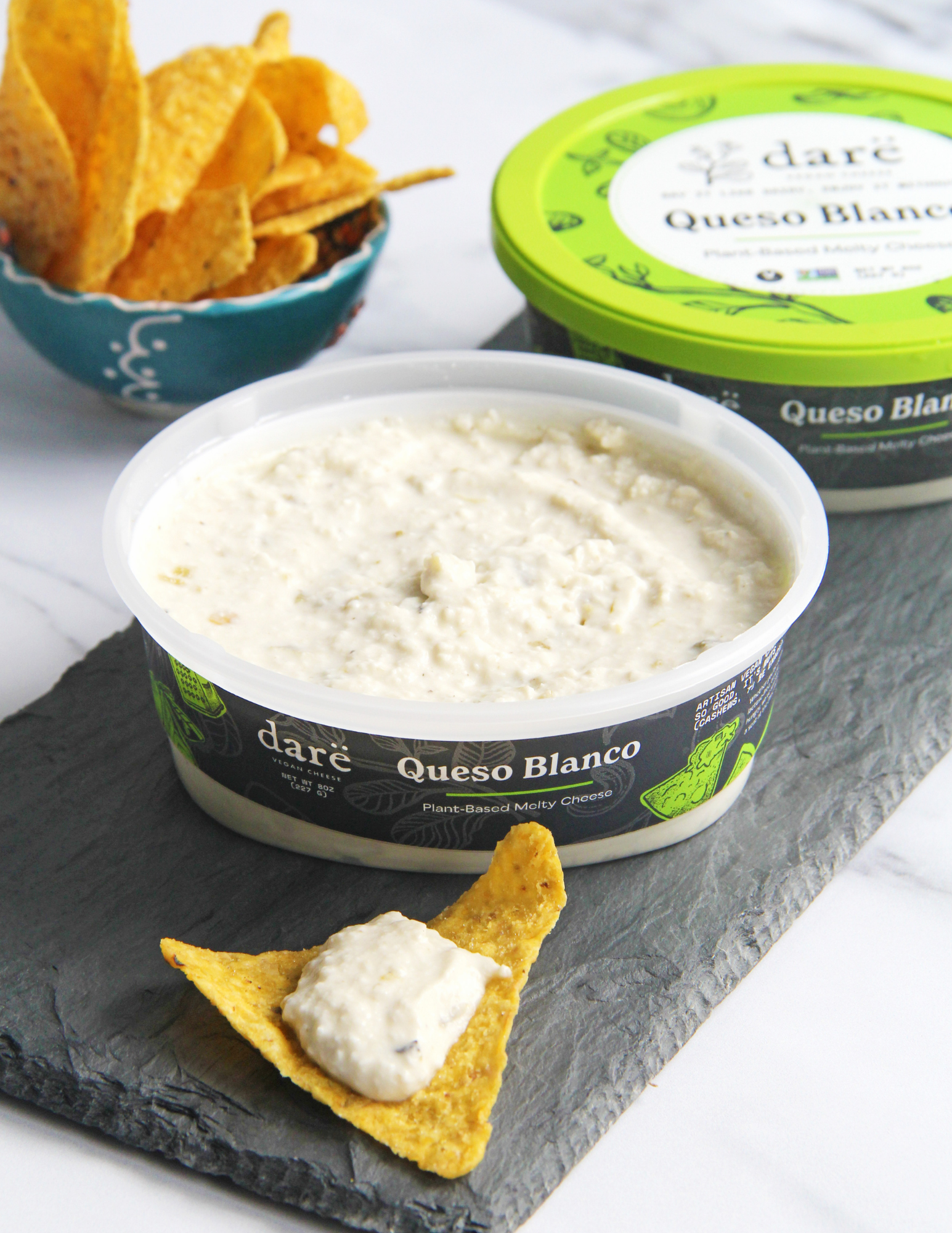 A tub of daré Queso Blanco plant-based cheese dip in the foreground on a dark slate board, with a tortilla chip dipped in the dip. A blue bowl of tortilla chips and another view of the green-lidded product container are blurred in the background.
