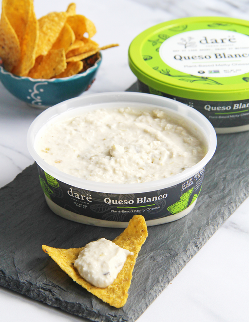 Load image 1 in gallery view. A tub of daré Queso Blanco plant-based cheese dip in the foreground on a dark slate board, with a tortilla chip dipped in the dip. A blue bowl of tortilla chips and another view of the green-lidded product container are blurred in the background.