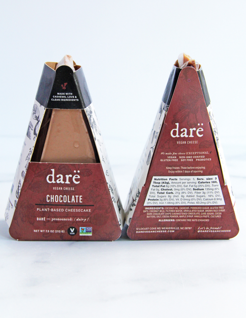 Load image 1 in gallery view. Two Daré Vegan Cheese chocolate cheese sticks in triangular cardboard packaging, shown side by side on a white marble surface against a white background. The left shows the brown cheesecake inside, while the right package shows the back with product details and nutritional information.
