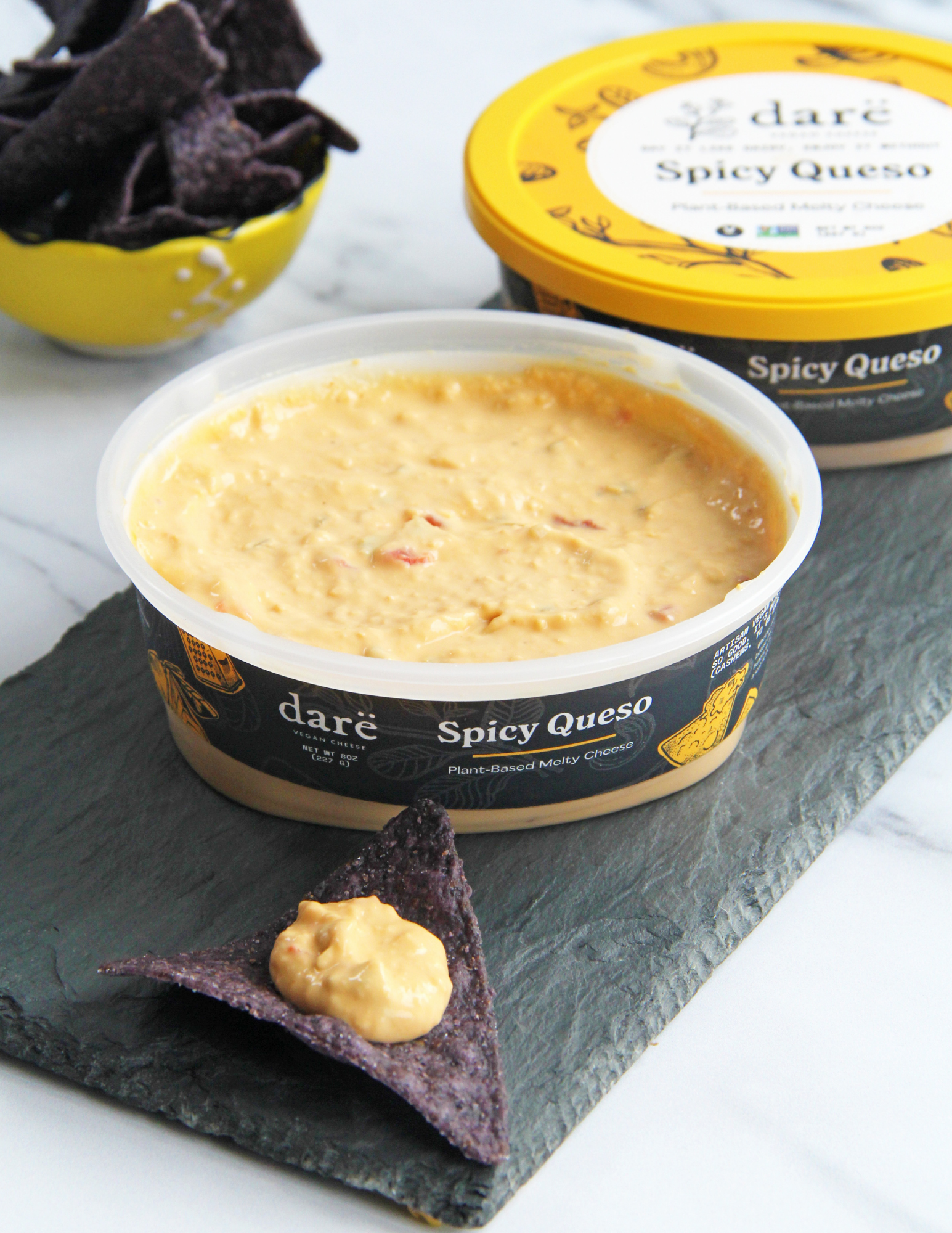 A tub of daré Spicy Queso dip in the foreground on a dark slate board, with a blue tortilla chip resting in front and a yellow tortilla chip in the background next to another tub of the product.
