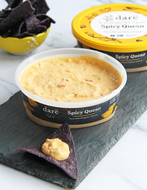 Load image 1 in gallery view. A tub of daré Spicy Queso dip in the foreground on a dark slate board, with a blue tortilla chip resting in front and a yellow tortilla chip in the background next to another tub of the product.