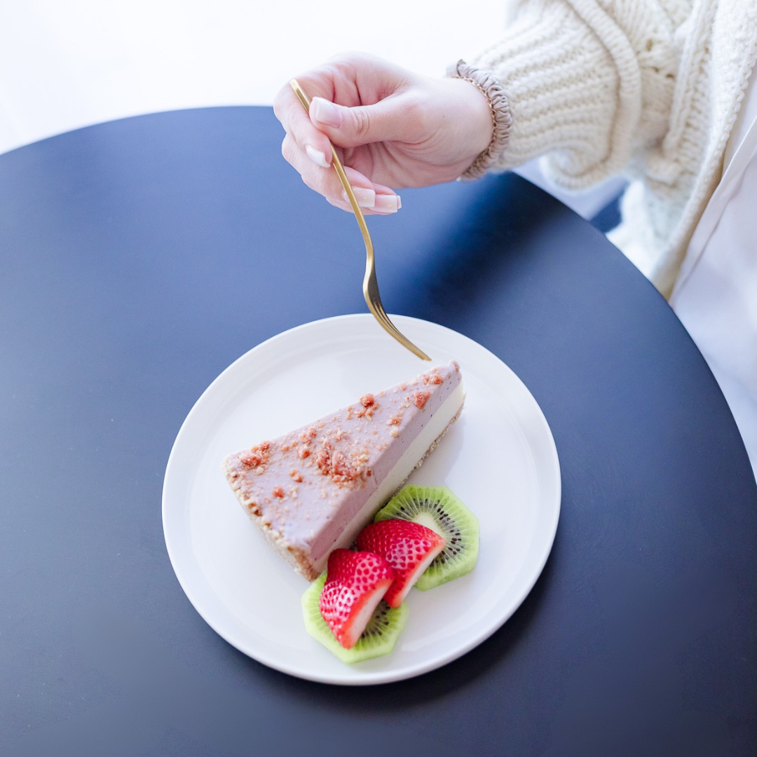 Darë Vegan Cheese Strawberry Shortcake Cheesecake on a plate with fresh fruit. hand holding gold fork hovering above, plate on blue circular table