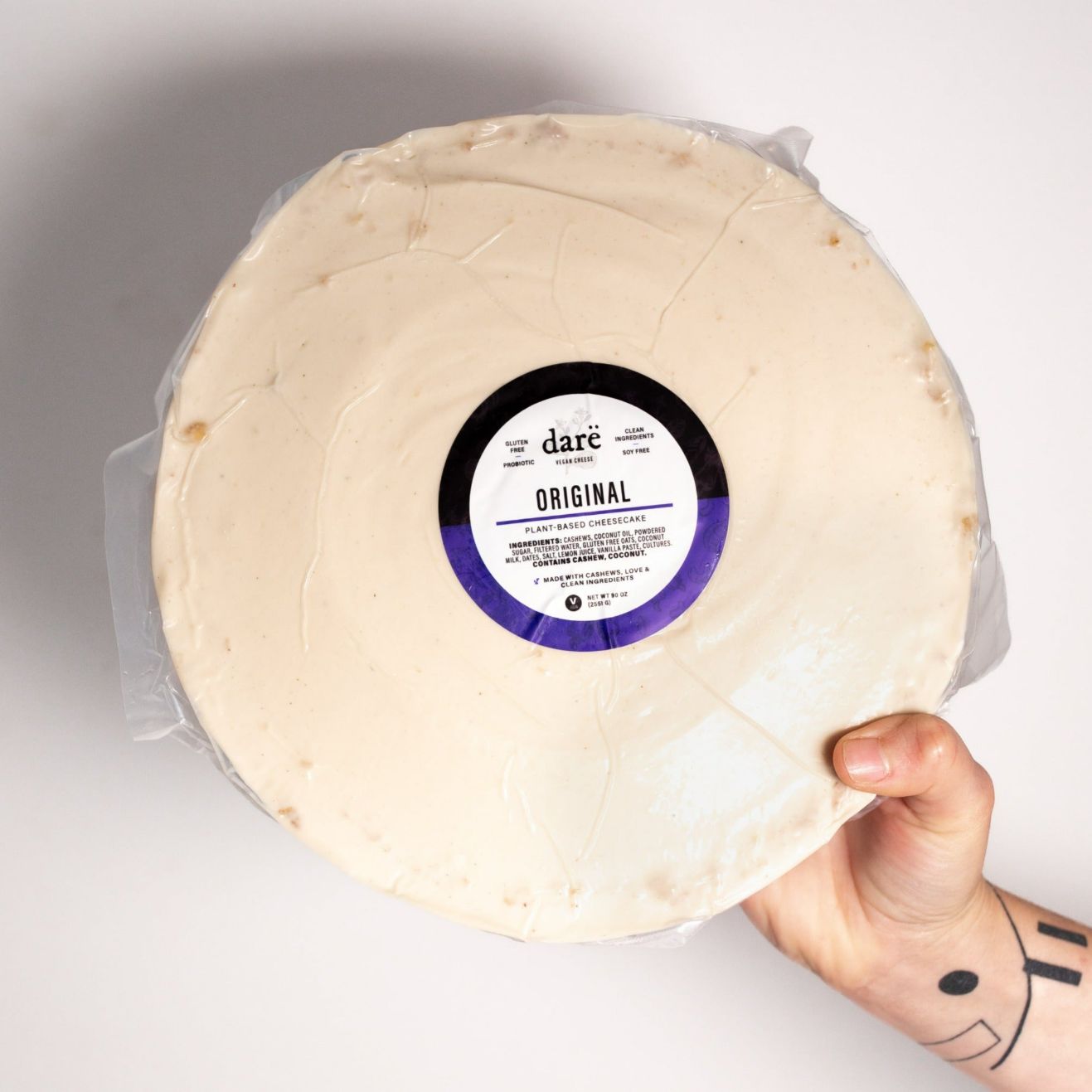 whole round vegan cheesecake being held by a human hand. label reads dare original plant-based cheesecake, wrapped in plastic.