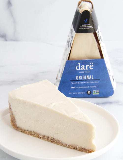 Load image 1 in gallery view. A slice of dare vegan cheese plant-based cheesecake with a gluten free vegan crust sits on a white plate, with the triangular blue and white package standing behind it on a light marble surface.