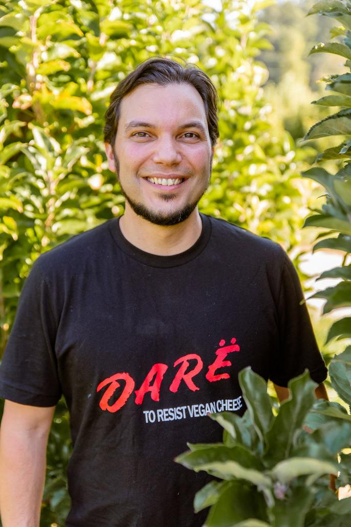 guy surrounded by nature with black tshirt on. red lettering reads dare white lettering reads to resist vegan cheese