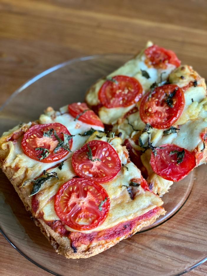 square pizza slices on a glass plate. topped with sliced tomato and basil shreads. made with vegan mozzarella cheese